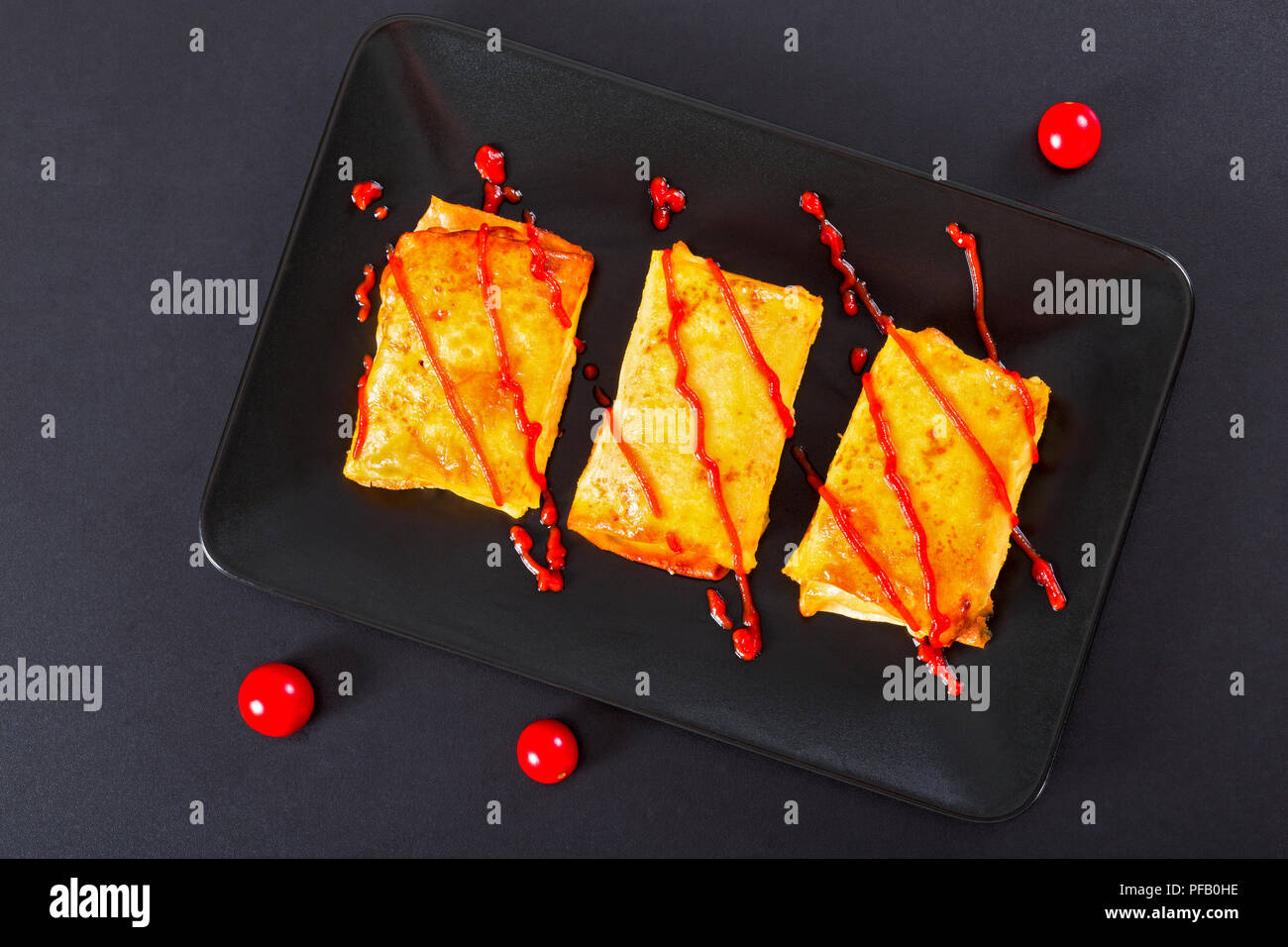 Rollen mit Pfannkuchen oder Pfannkuchen mit Hackfleisch mit Ketchup auf Teller mit Ketchup gegossen gegossen gefüllte Kirschtomaten auf schwarzen Hintergrund, Ansicht von ab Stockfoto