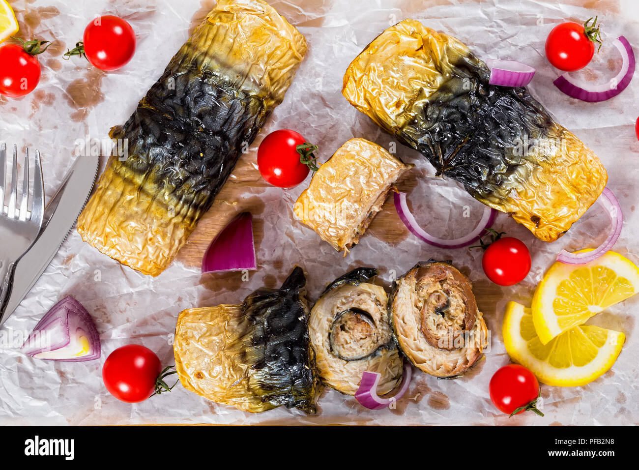 Gesunde gebackenes Filet Makrelen in Rollen auf weißem Pergamentpapier mit Messer und Gabel, Zwiebel, Tomaten, und Zitronenscheiben auf Hintergrund, Ansicht von oben Stockfoto