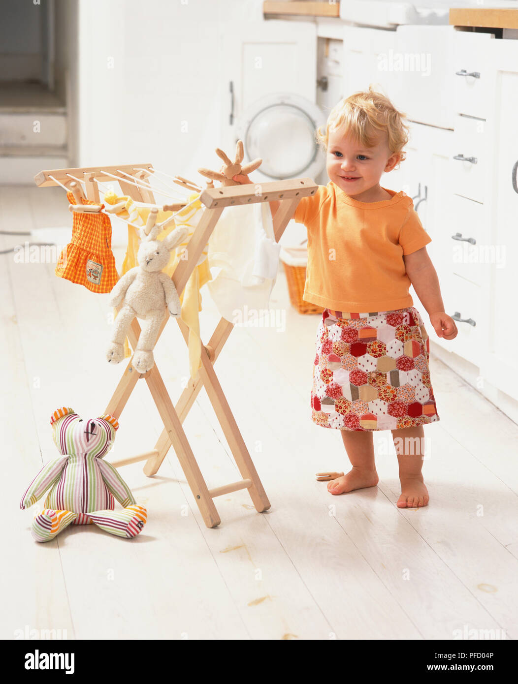 Lächelnd blonden Kleinkind in gelben T-Shirt und Schürze in der Küche hängenden Kleidungsstücken und weiches Spielzeug auf der Wäscheleine, Vorderansicht. Stockfoto