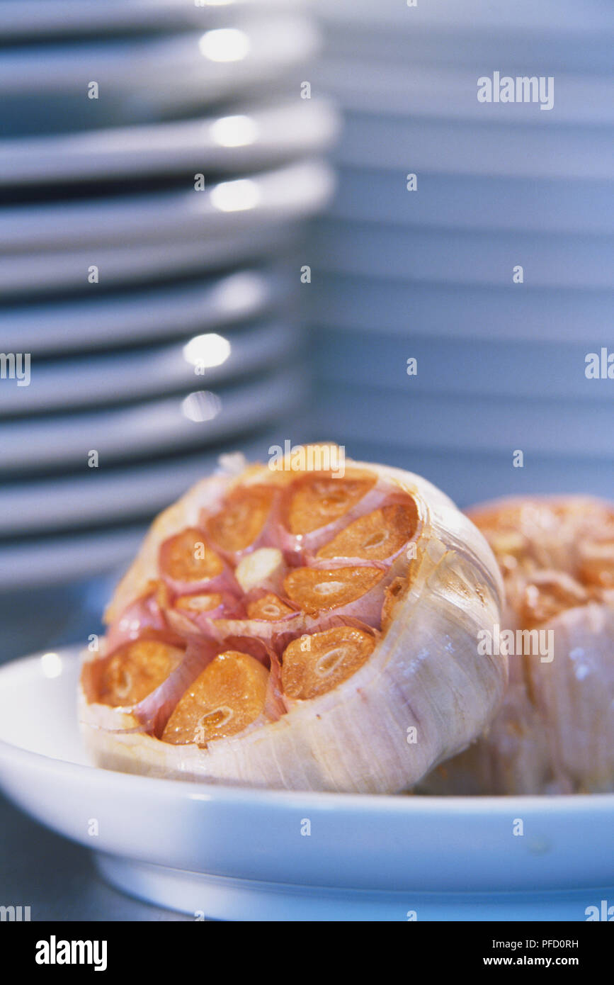 Allium Sativum, Knoblauch, zwei gerösteten Knoblauch Zwiebeln, Tops abgeschnitten, auf einem weißen Teller angeordnet Stockfoto