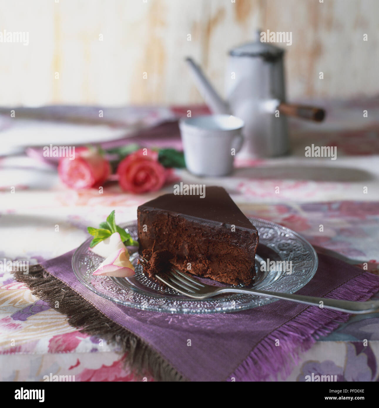 Stück Schokolade Kuchen auf Glasplatte, neben Silber Gabel und dekorativen rose bud, auf lila Unterlage, Rosenknospen, Teekanne und Tasse im Hintergrund, Tischdecke mit Blumenmuster Stockfoto
