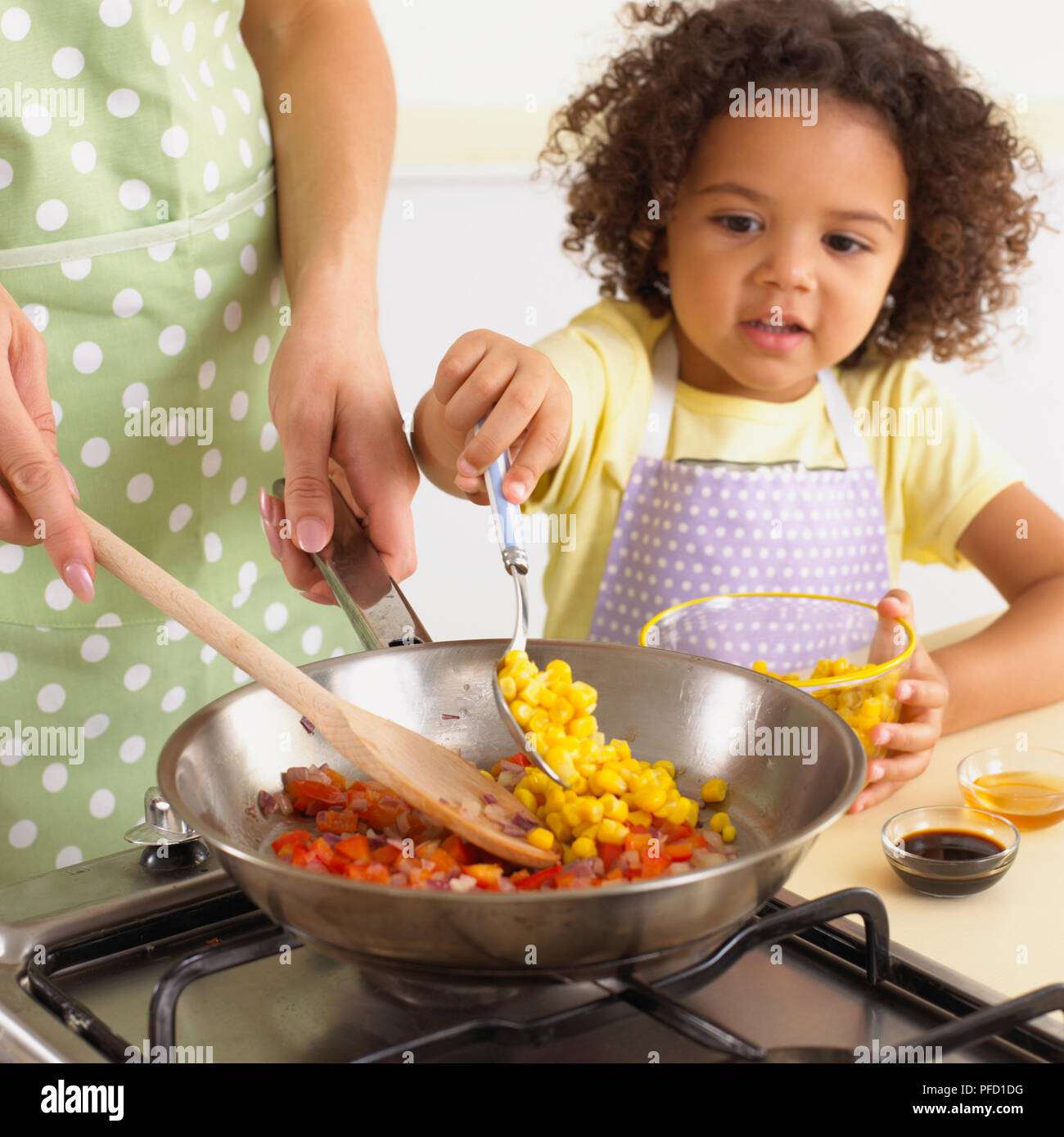 Mädchen hinzufügen Zuckermais mit Löffel, neben Frau rühren, Zwiebeln und Paprika in der Pfanne, Schalen von Zuckermais, Essig und Honig auf der Küchenarbeitsplatte, close-up Stockfoto
