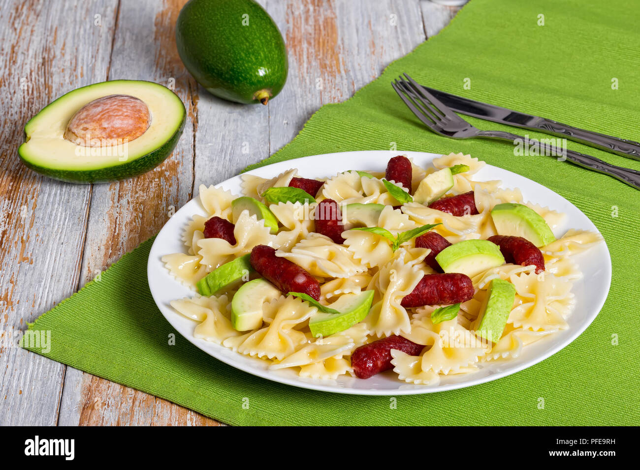 Mini Salami farfalle Pasta warmer Salat mit Avocado Scheiben und Blätter Basilikum auf weißen Teller am Tisch mit Messer und Gabel, die Hälfte der reife Avocado auf ol Stockfoto