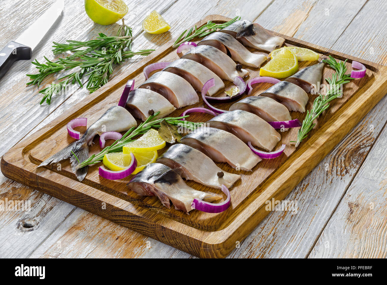 Marinierte Fischfilets frische Makrele Fisch in Scheiben geschnitten auf Schneidebrett mit Rosmarin, Zitrone, roten Zwiebeln und Gewürzen, Messer und Zitrone Stücke o Stockfoto