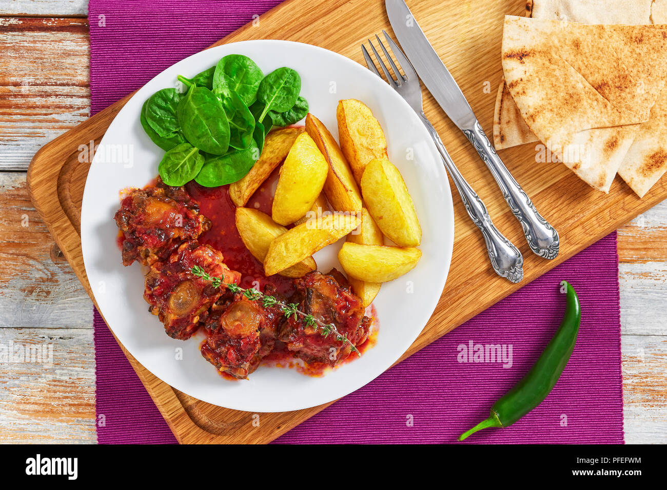 Rabo de Toro oder ochsenschwanz Eintopf Serviert mit Spinat, Salat und gebratene Kartoffel auf weiße Platte auf Schneidebrett mit Gabel, Messer und pita Brot. jalapeno Pepp Stockfoto