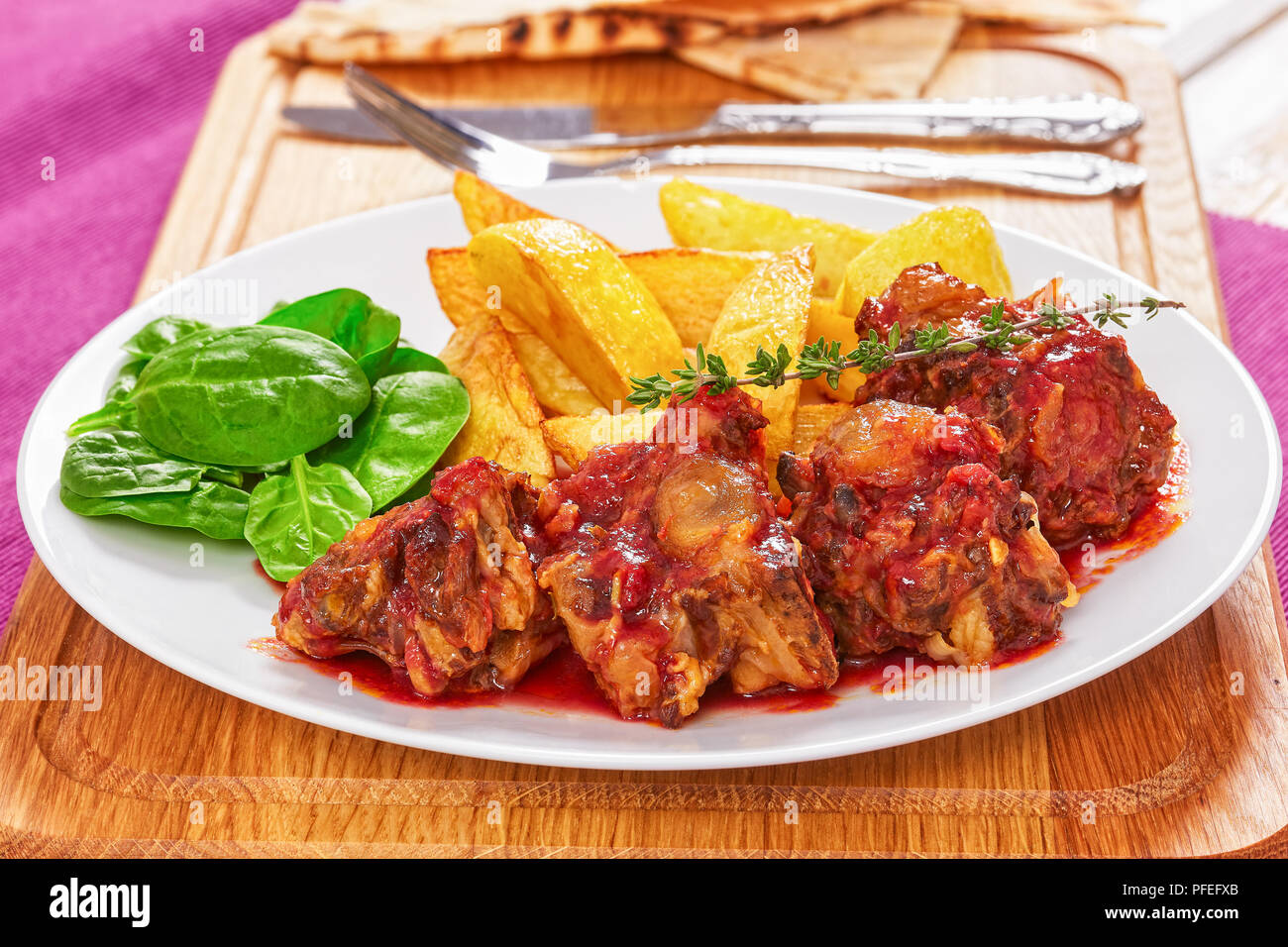 Ochsenschwanz in Rotwein geschmort mit Spinat Salat und gebratene Kartoffel auf weiße Platte auf Schneidebrett mit Gabel, Messer und pita Brot. jalapeno Pfeffer Stockfoto