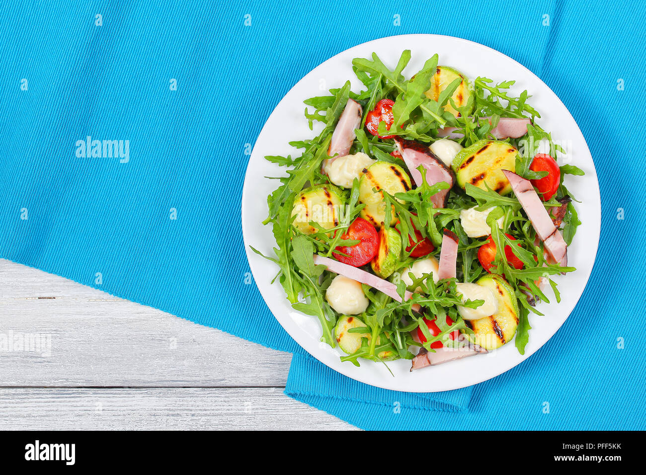 Köstliche gegrillte Zucchini, Mozzarella, Käse, Fleisch, Tomaten und Rucola Sommer Salat auf weißen Teller am Tisch Matte, Ansicht von oben Stockfoto