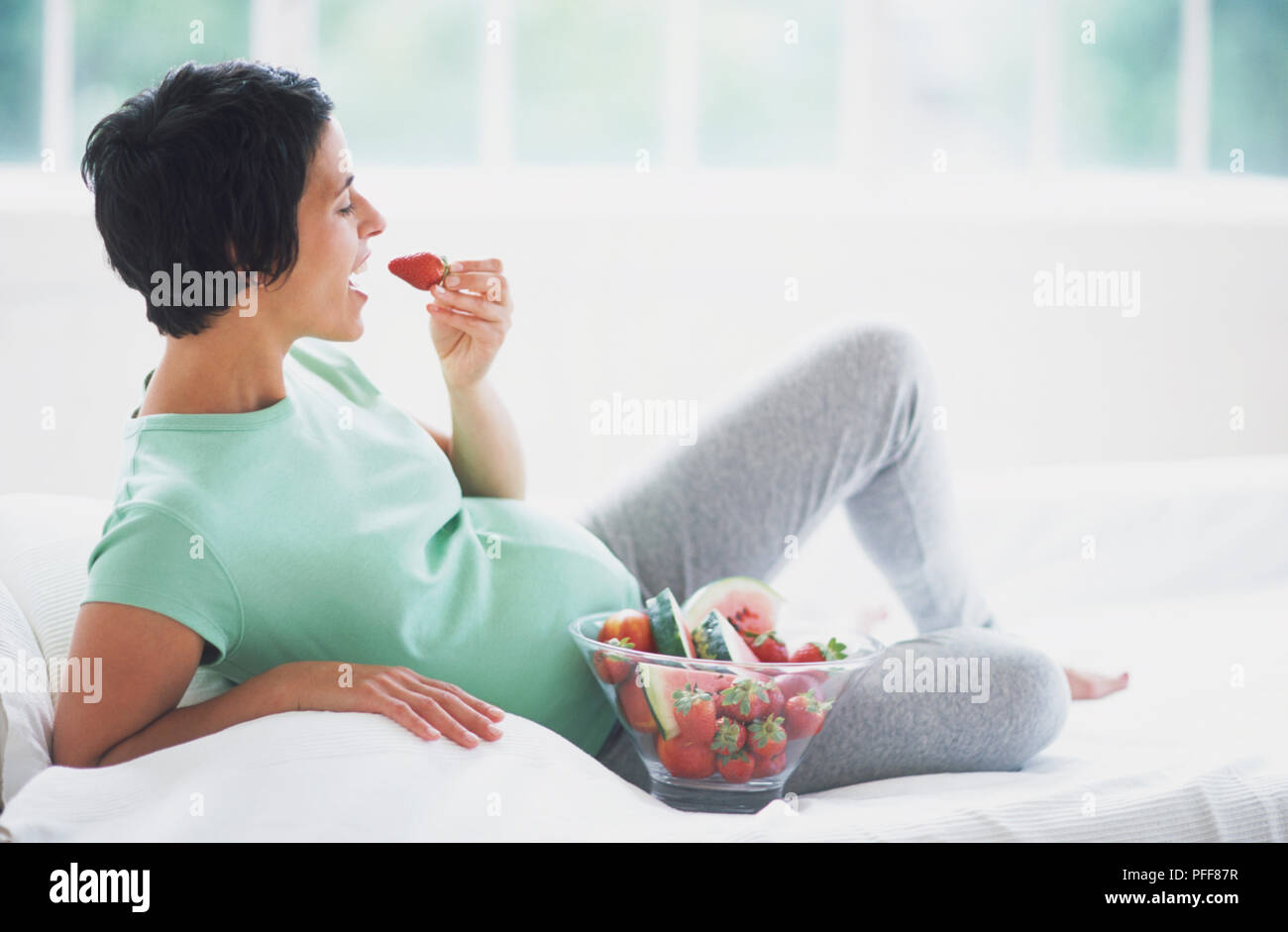 Frau in frühen Stadien der Schwangerschaft liegen auf dem Sofa und essen Erdbeeren aus Glas Schüssel, Seitenansicht. Stockfoto