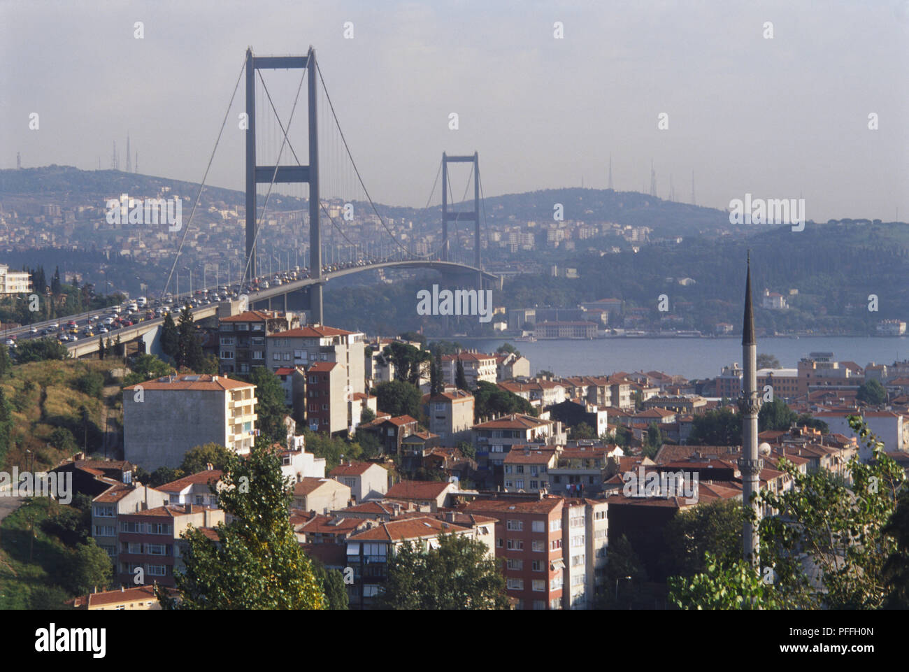 Asien, Türkei, Istanbul, den Bosphorus Hängebrücke überspannt den Fluss zwischen Ortakoy und Beylerbeyi, Stadtbild und Minarett im Vordergrund, viele Fahrzeuge über die Brücke fahren, moderne Gebäude der opposide Bank eingehüllt in Nebel im Hintergrund. Stockfoto
