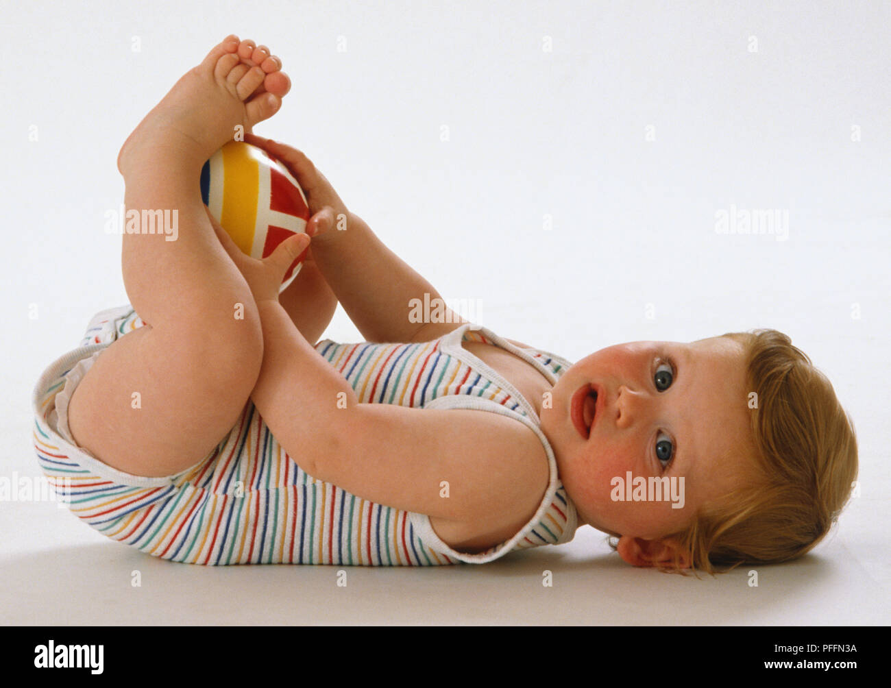 Baby tragen gestreifte Jumpsuit, liegend auf seinem Rücken, umklammerte bunten Ball zwischen Händen und Füßen. Stockfoto