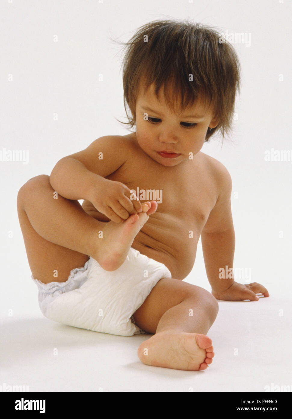 Baby tragen Windeln sitzen, ein Bein in der Nähe seiner Brust gezogen, berührend und prüft seine Zehen. Stockfoto