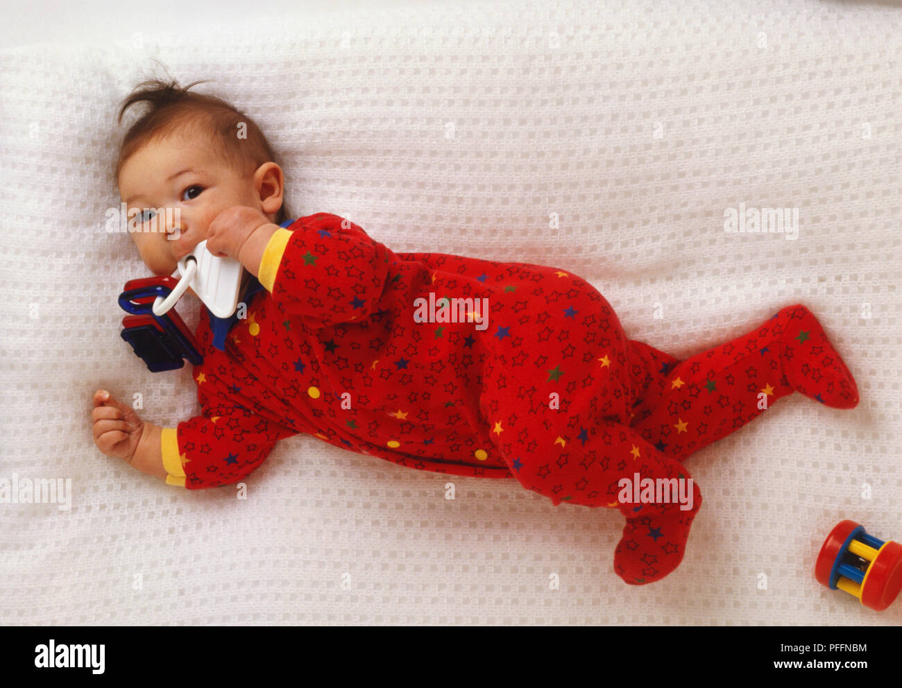Baby das Tragen der roten Overall, Haare stehen am Ende, der lag auf der Seite und auf Bett, Saugen aus Kunststoff Spielzeug Schlüsselanhänger, oben betrachten. Stockfoto