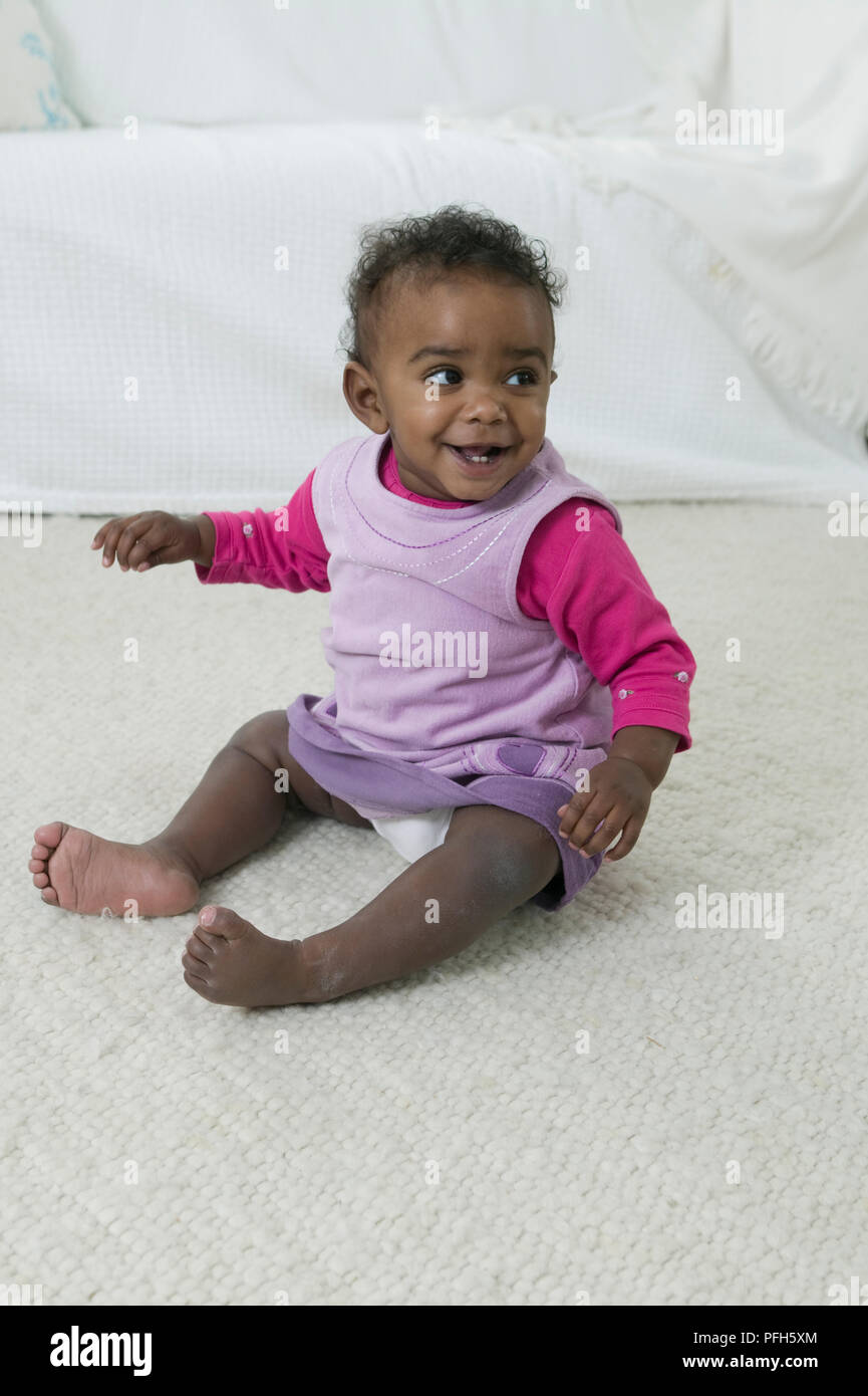 Baby Mädchen in lila und rosa Kleidung sitzen auf dem Boden, lächelnd, Suchen nach Stockfoto