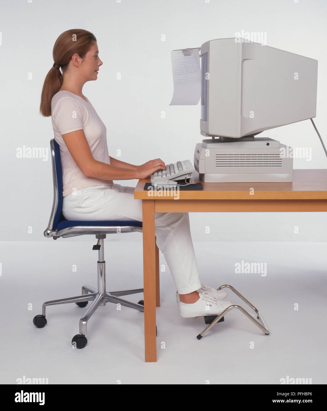 Frau sitzt an einem Schreibtisch mit einem Computer, Füße auf eine Fußstütze, Seitenansicht Stockfoto
