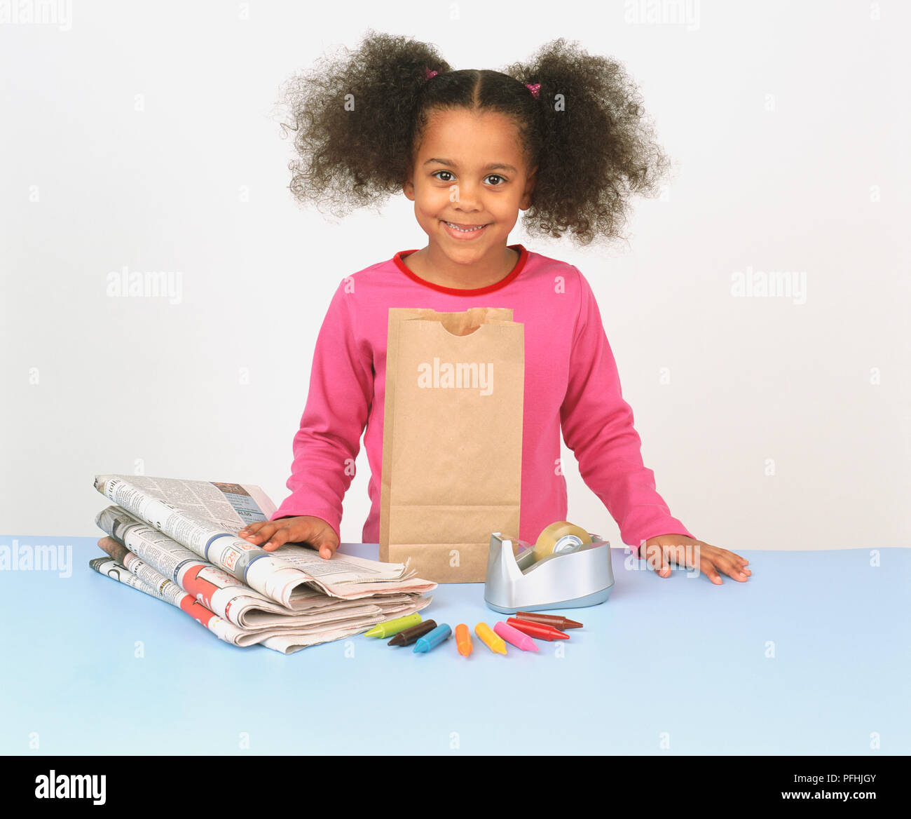 Mädchen mit Pigtails, lächelnd, hinter einem blauen Schreibtisch mit einer Papiertüte, sticky tape Dispenser, Zeichenstifte, Zeitung. Stockfoto