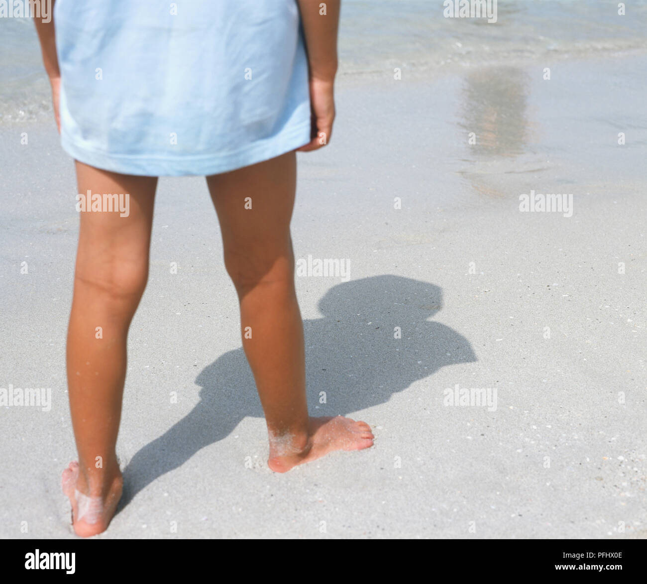 Gebräunte Beine der Person tragen lange T-Shirt am Strand Wasser - Kante mit Schatten fallen auf Sand, Vorderansicht. Stockfoto