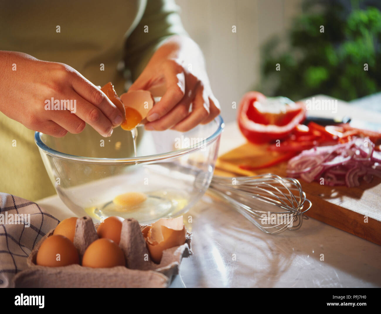 Rohes Ei tropft vom Shell über Glasschale, ei Karton und schneebesen von Seiten der Schüssel eingebrochen wird, gehackte Gemüse auf Holzbrett im Hintergrund. Stockfoto