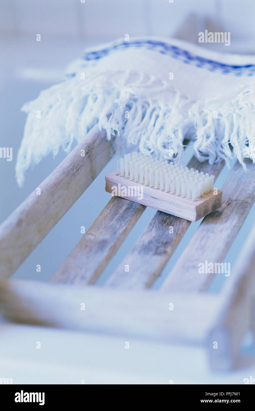 Schrubber mit kurzen gelben Borsten und Holzgriff, ruht auf Holz- Fach der gleichen Farbe, der Bretter mit Lücken dazwischen, gefalteten weißen Teppich mit dekorativen blauen Streifen und Quasten im Hintergrund Stockfoto