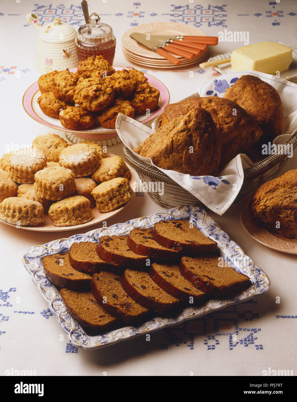 Anzeige von Muffins, Scones, Scheiben von Schokolade Kuchen und crusty, keilförmige Brötchen, Honig, Butter, Teller und Messer, auf Bestickten weißen Tischdecke Stockfoto