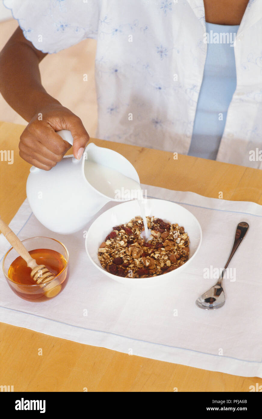 Gießen Milch von Kanne auf Müsli in der Schüssel, daneben eine Schale mit Honig und einem Löffel heraus auf weißer Unterlage gelegt Stockfoto