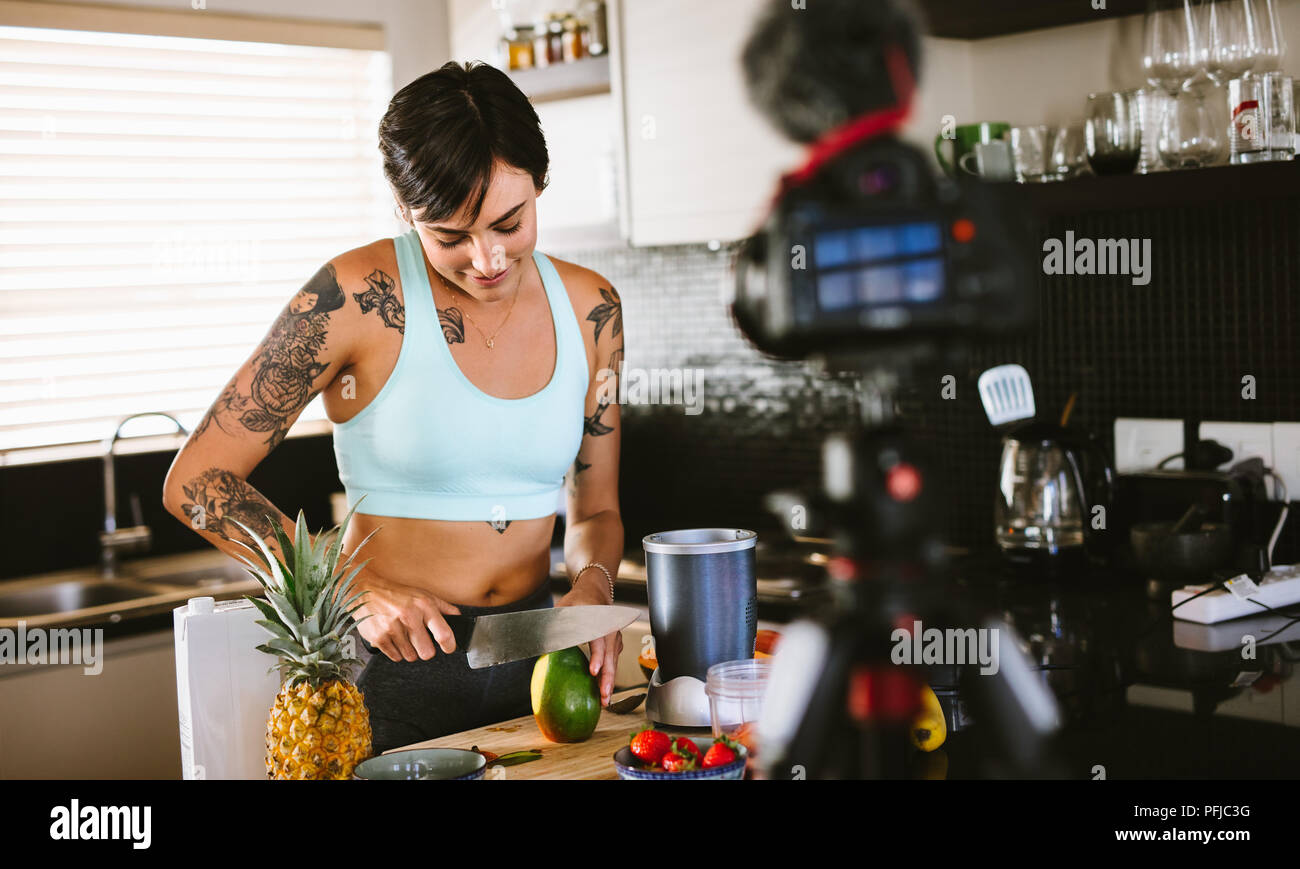 Tätowierte junge Frau schneiden Obst auf der Arbeitsplatte in der Küche und der Aufnahme von Video auf der Kamera. Social media Einflussnehmer in Küche, Smoothies und PCI Stockfoto