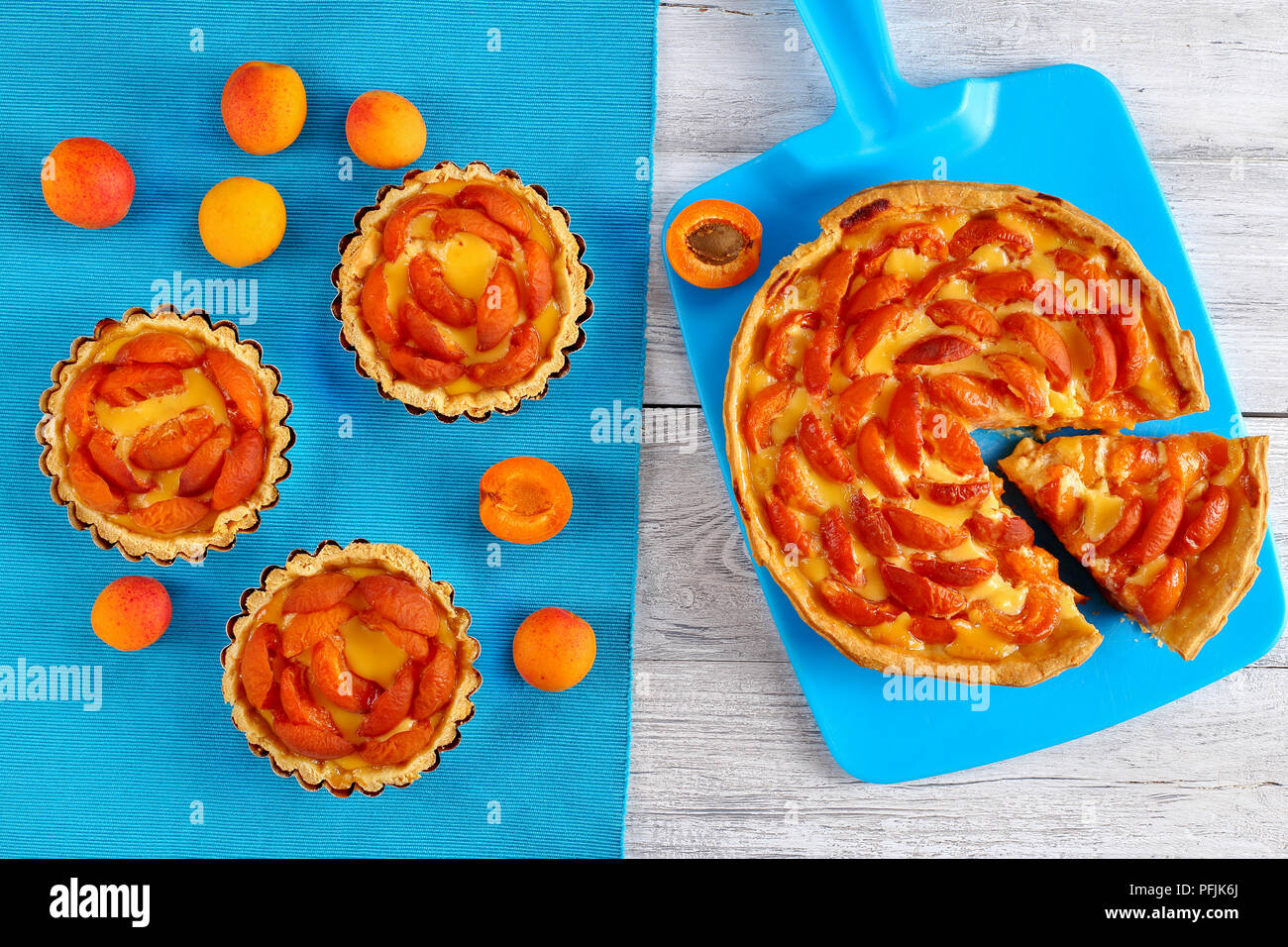 Leckere hausgemachte Aprikosen Kuchen mit Vanillepudding Creme mit einem Stück beiseite auf Schneidebrett und drei Torten mit reifen Aprikosen auf Tisch matte Hori Stockfoto
