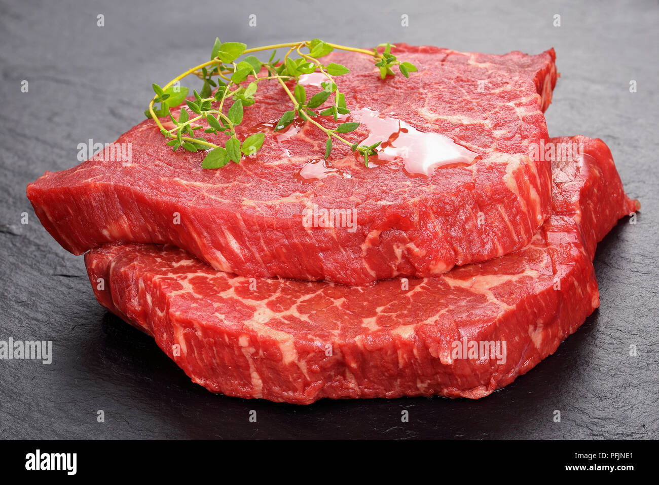 Zwei Marmorierte Rindfleisch steaks Ausgegossen über mit Olivenöl und garniert mit frischem Grün Thymian auf schwarzem Schiefer Fach, Ansicht von oben, close-up Stockfoto