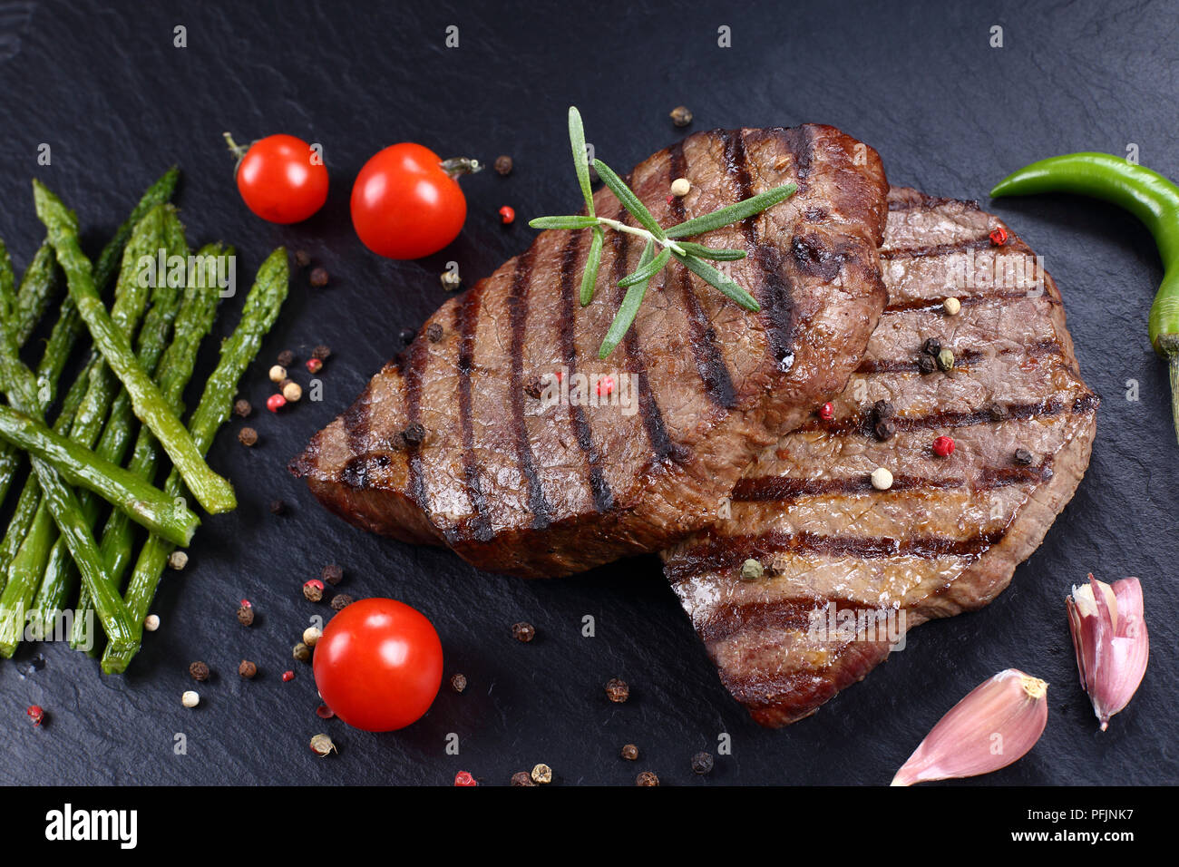 Hot köstliche gegrillte Rindersteaks mit Streifen mit Tomaten, gegrillter Spargel, Tomaten, Knoblauch und Gewürzen auf schwarzem Schiefer Teller serviert, horizontal Stockfoto