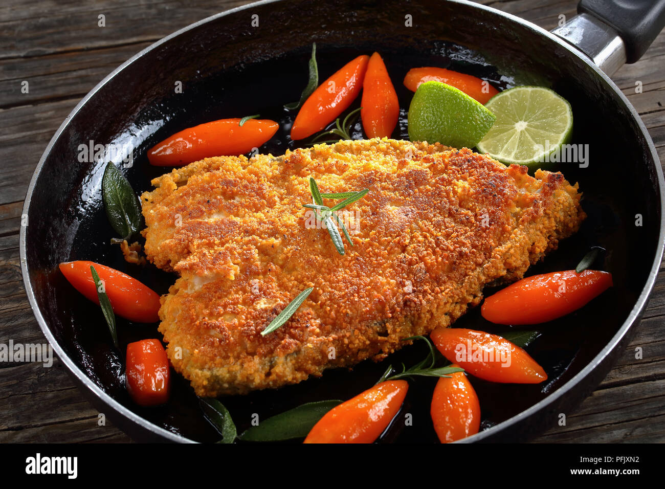 Gebratene panierte Steaks von Meer Fisch in der Pfanne mit glasierten Karotten, schneiden Sie in Tourne (ovale Form), frische Kalk Schichten und aromatischen Kräutern, horizontale Ansicht her Stockfoto
