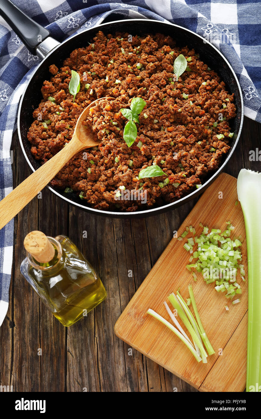 Leckere bolognese Ragout-Zutat für Pasta oder Lasagne in der Pfanne auf Holztisch mit Löffel, Handtuch und Schneidbrett mit fein gehackten Cel Stockfoto