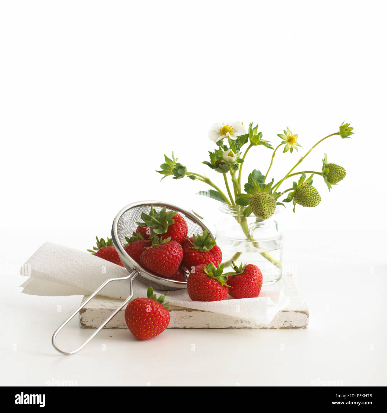 Reife rote Erdbeeren im Sieb, neben Stämme der Unreife Erdbeeren und Blumen im Glas Stockfoto