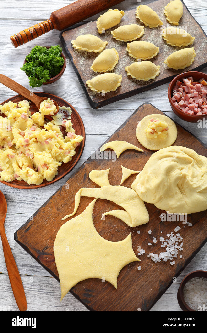 Ungekocht pierogi - Polnische und ukrainische Knödel. Kartoffelpüree, fein gehacktem Schinken oder Speck in Schalen, Teig auf Holz Schneidebrett und Ro Stockfoto