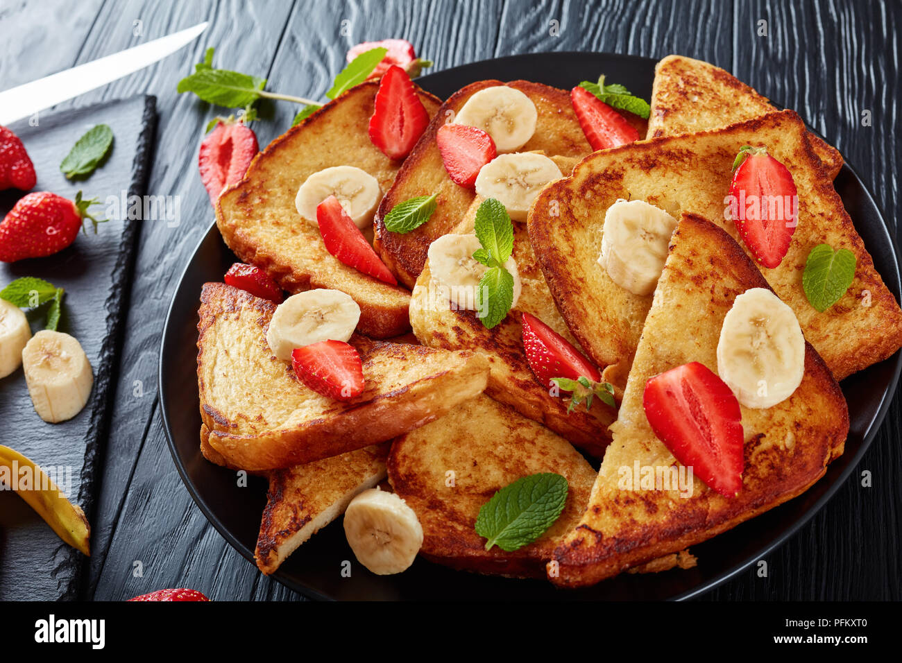 French Toast mit appetitlich goldbraune Kruste und flauschige Innenseite serviert auf einem Teller mit frischen Erdbeeren, Bananenscheiben, Minze. Zutaten auf Schnitt Stockfoto