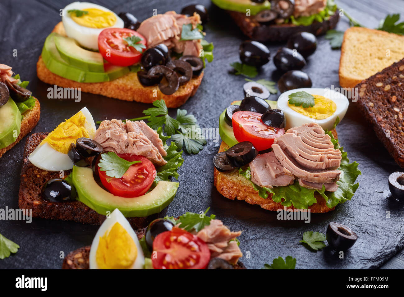 In der Nähe von Thun Sandwiches mit Avocado, Salat, Tomaten, schwarze Oliven und hart gekochte Ei auf Roggen und Mais getoasteten Brotscheiben auf einem schwarzen s Stockfoto