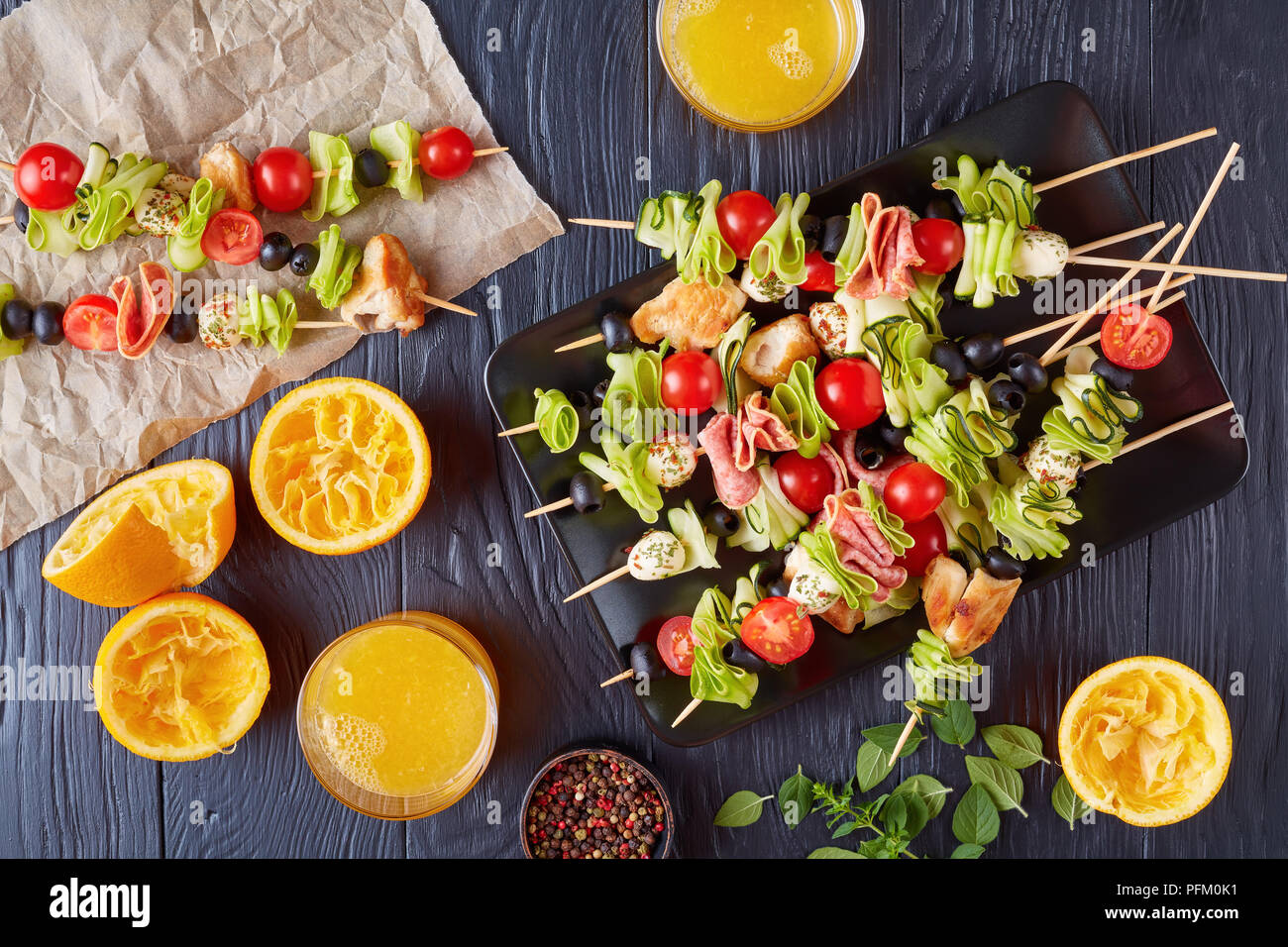 Antipasti Spieße mit Hühnerfleisch, rohe Zucchini, Tomaten, Mozzarella Kugeln, Scheiben Salami, Oliven auf einem Schild an einem Holztisch mit Orange Stockfoto