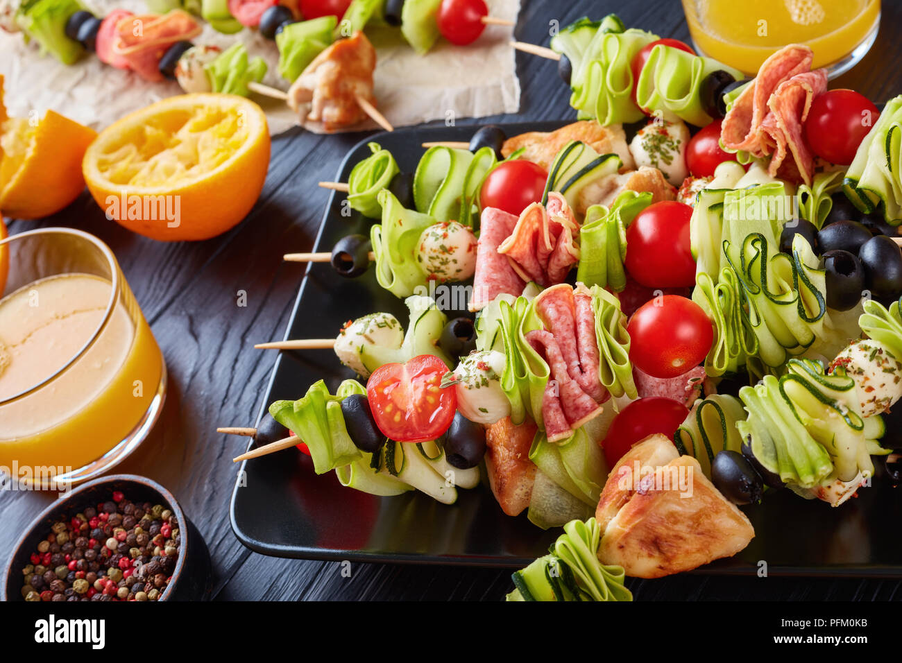 Spiesse mit Hähnchenfleisch, Zucchini, Tomaten, Mozzarella Kugeln, Scheiben  Salami, Oliven auf einer schwarzen Platte auf einem Holztisch mit  Orangensaft in Glas c Stockfotografie - Alamy