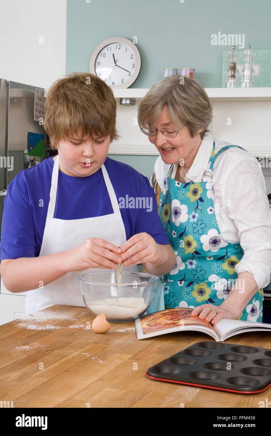 Junge cracking Egg über Schüssel, ältere Frau neben ihm beobachten, Kochbuch und törtchenform auf Arbeitsfläche Stockfoto