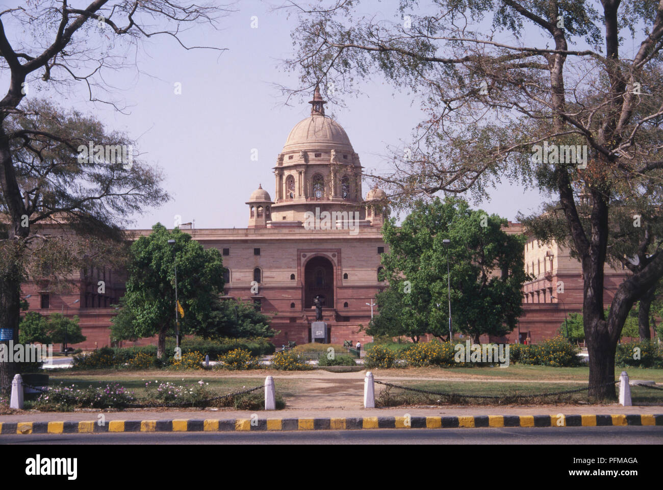 Indien, Delhi, Vijay Chowk, South Block Gehäuse Amt des Ministerpräsidenten und das Verteidigungsministerium, großes rotes Gebäude, hohe Kuppel, Statue im Innenhof, Bäume, gestreifte Vorfahrt im Vordergrund. Stockfoto
