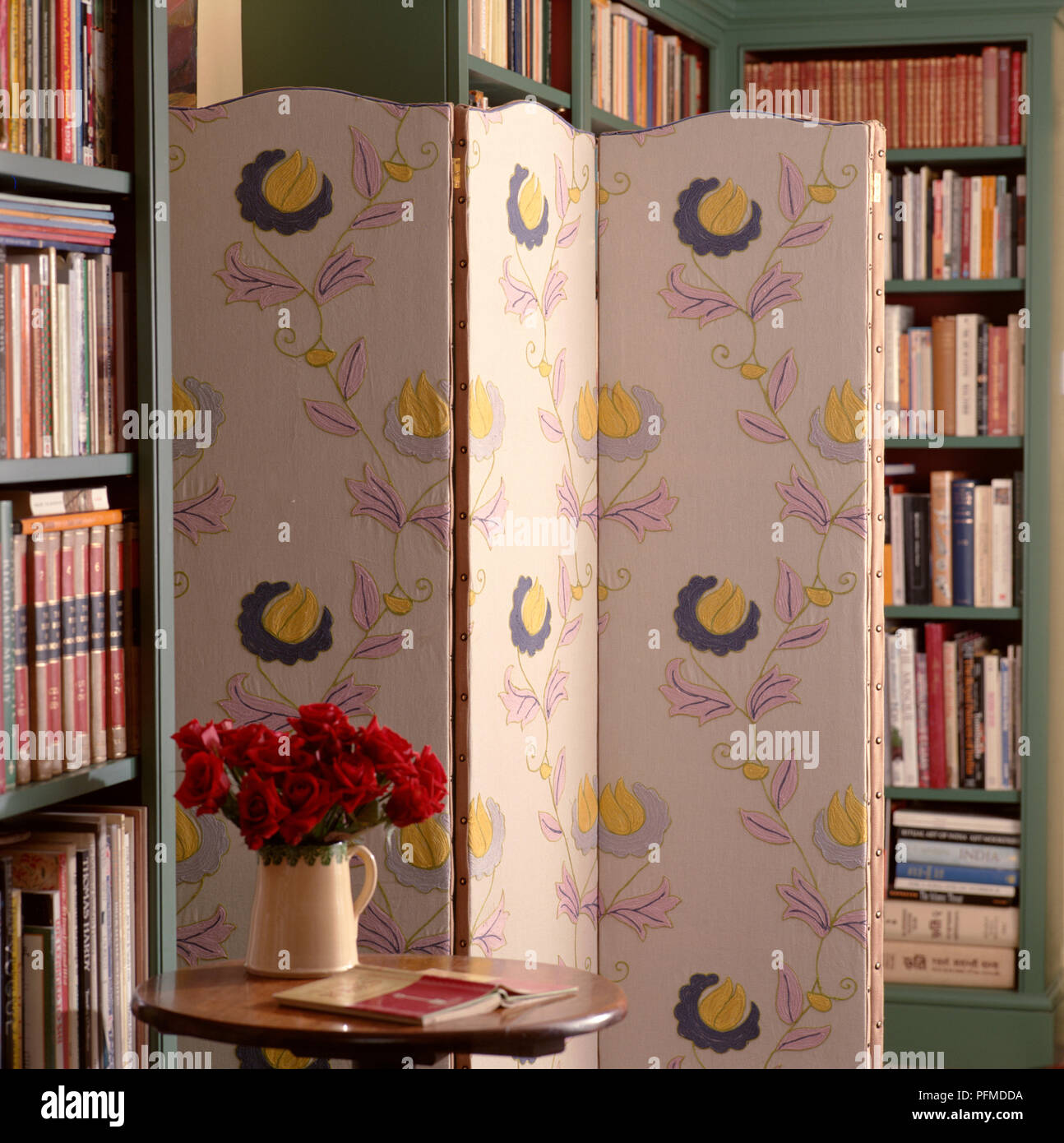 Gemusterten Stoff Bildschirm stehen im Zimmer voller Bücher, Holztisch mit einer Vase mit Rosen im Vordergrund, hohen Bücherregale im Hintergrund. Stockfoto