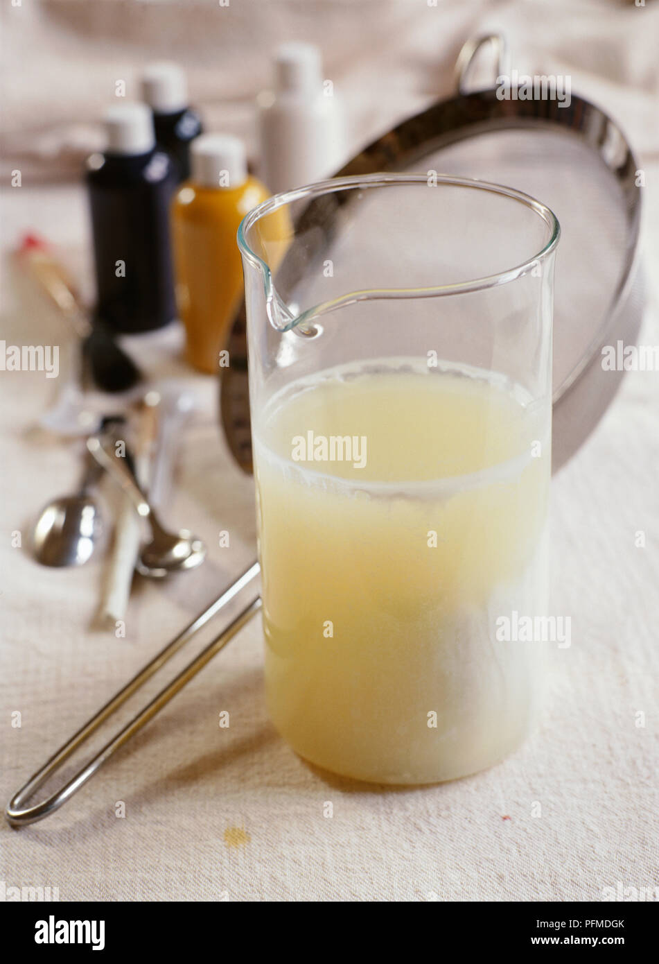 Glas Krug Milch Quark und Molke Milch Paint zu machen, hellgelbe Flüssigkeit mit weißem Quark, auf weiße Tischtuch; Metallsieb, kleine Farbtöpfe und Teelöffel im Hintergrund. Stockfoto