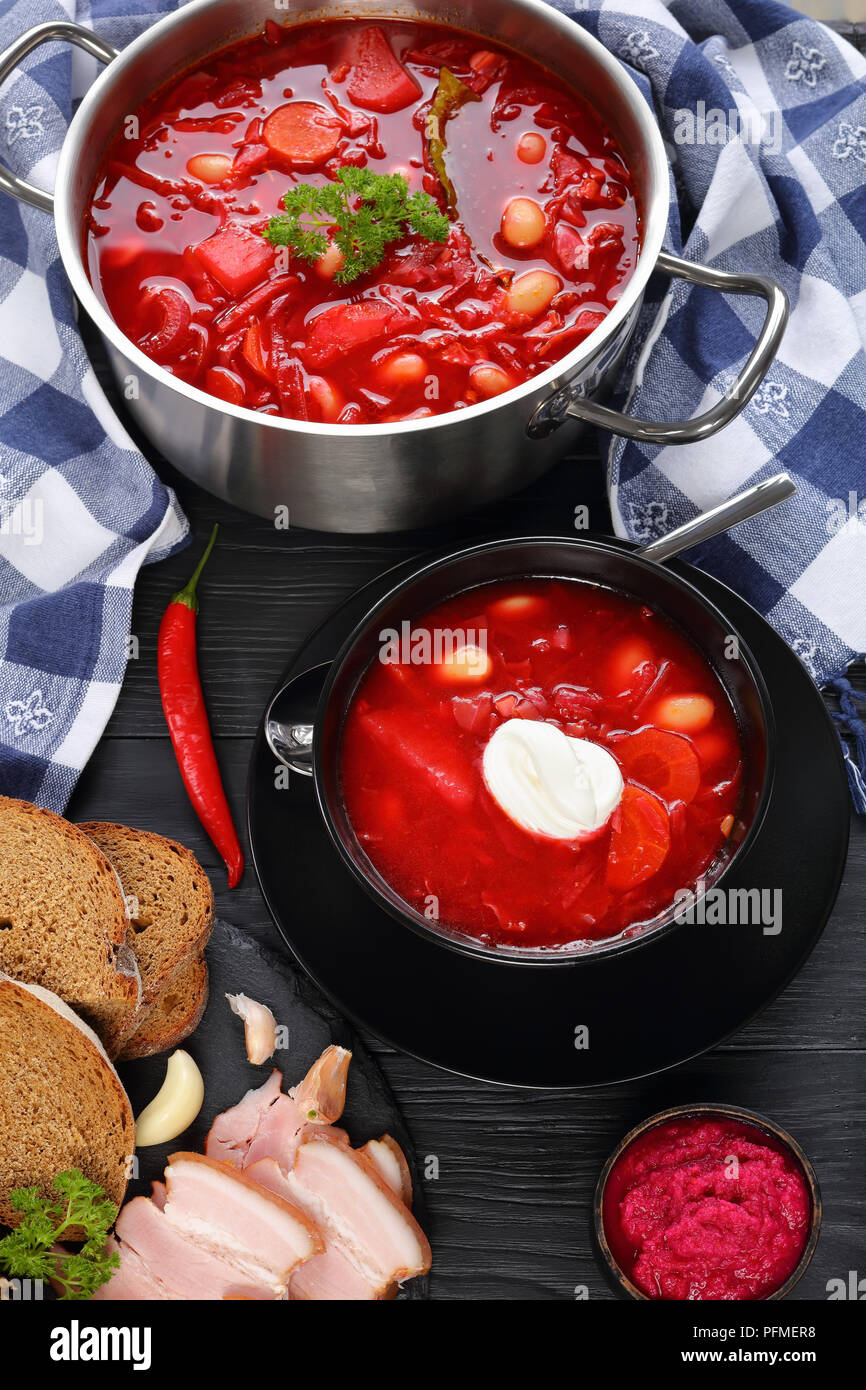 Köstliche rote Beete rote Suppe mit weißen Bohnen oder Borschtsch in Edelstahl Kasserolle schwenken und in Schwarz Schüssel mit Roggenbrot und geräuchertem Speck auf Tisch, Stockfoto