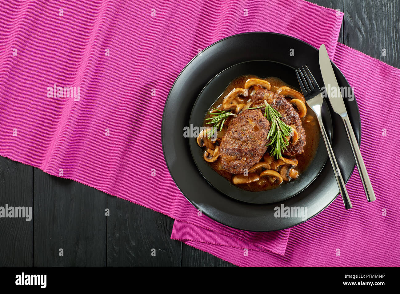 Teil des heißen herzhafte Steaks saftige Salisbury mit köstlichen Pilz Zwiebel Sauce auf schwarzem Teller mit Besteck serviert, Restaurant Stil, Blick von abov Stockfoto