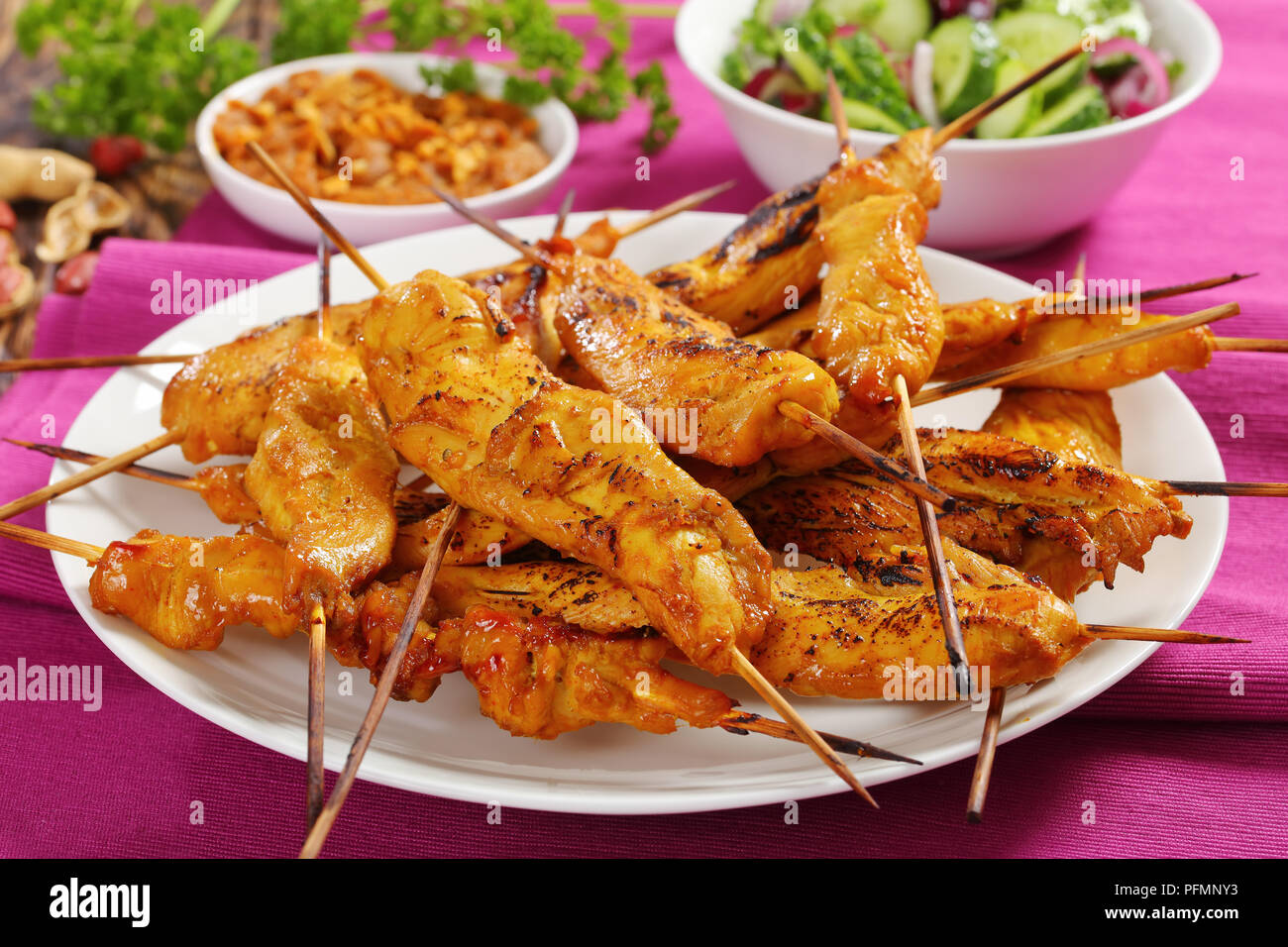 Close-up Chicken Satay auf Spieße auf einem weißen Teller auf rustikalen Holztisch mit Erdnusssoße und Gurke Zwiebel Salat serviert, authentische Rezept, Anzeigen Stockfoto