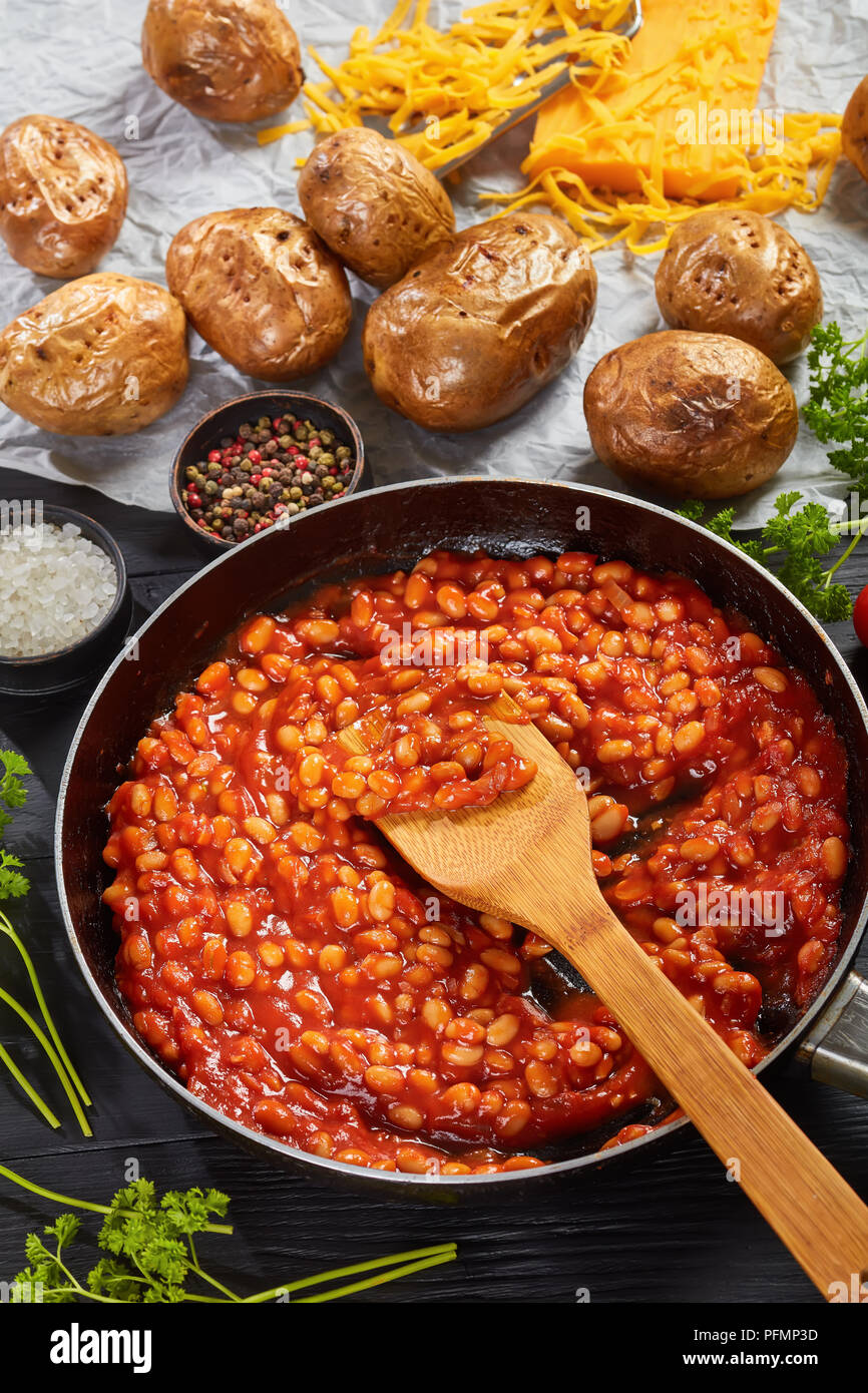 Gebackene Bohnen mit Tomatensauce in einer Pfanne. gebackene Kartoffel oder Pellkartoffeln mit Goldgelb knusprige Haut auf einem Papier mit Cheddar Käse im Hintergrund, Stockfoto