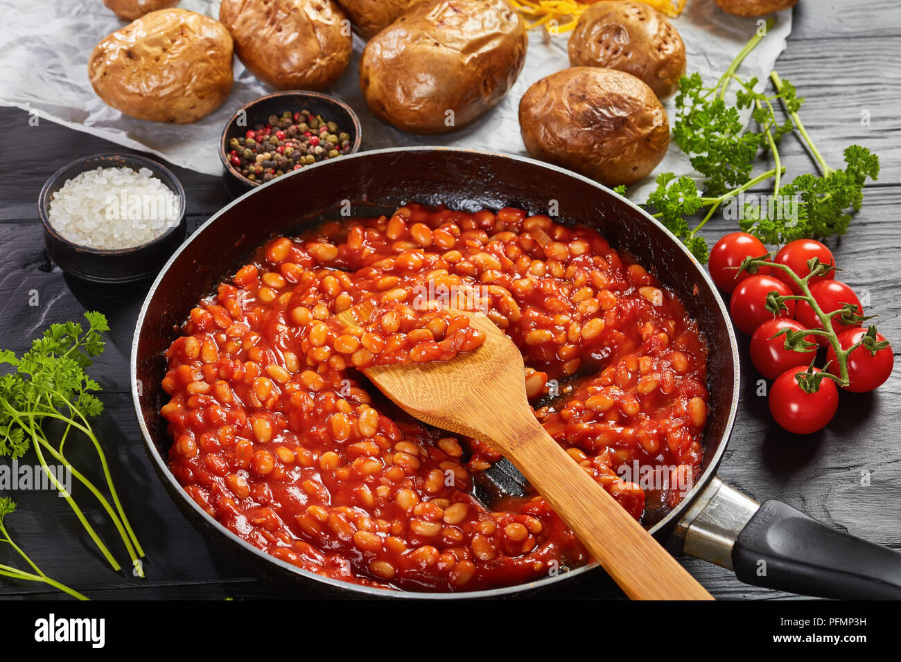 Bohnen mit Tomatensauce in einer Pfanne. gebackene Kartoffel oder Pellkartoffeln mit Goldgelb knusprige Haut auf einem Papier mit Cheddar Käse im Hintergrund, Verti Stockfoto