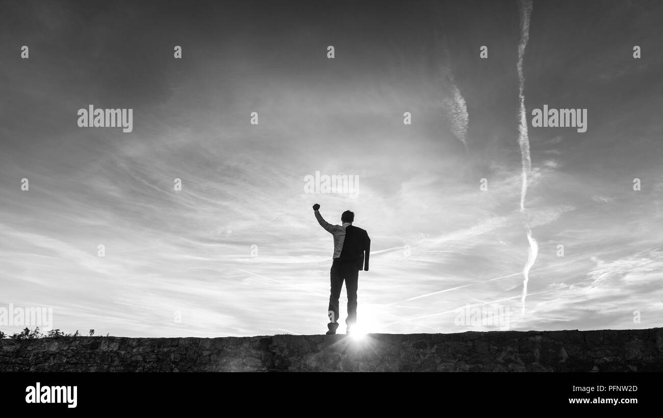 Ansicht der Rückseite des erfolgreichen Geschäftsmann mit seiner Faust erhoben, als er an einer Wand mit Hintergrundbeleuchtung durch eine Sunburst von der Sonne in einem konzeptionellen Graustufenbild steht. Stockfoto