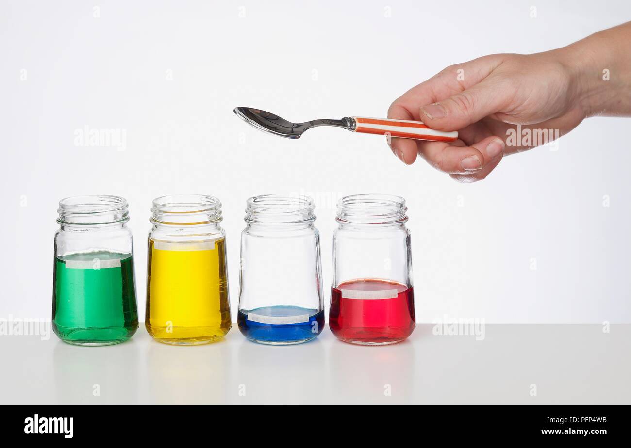 Person, die Löffel über vier Gläsern mit grünen, gelben, blauen und roten Flüssigkeiten Stockfoto