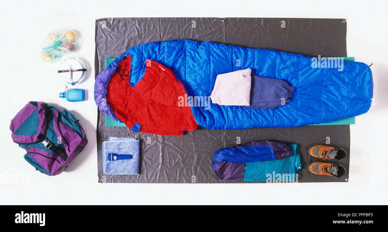 Blau und Rot Schlafsack auf einer Matte, die von Paar Schuhe, Kleidung, Rucksack, Essen und Trinken, Taschenlampe und Handtuch umgeben Stockfoto