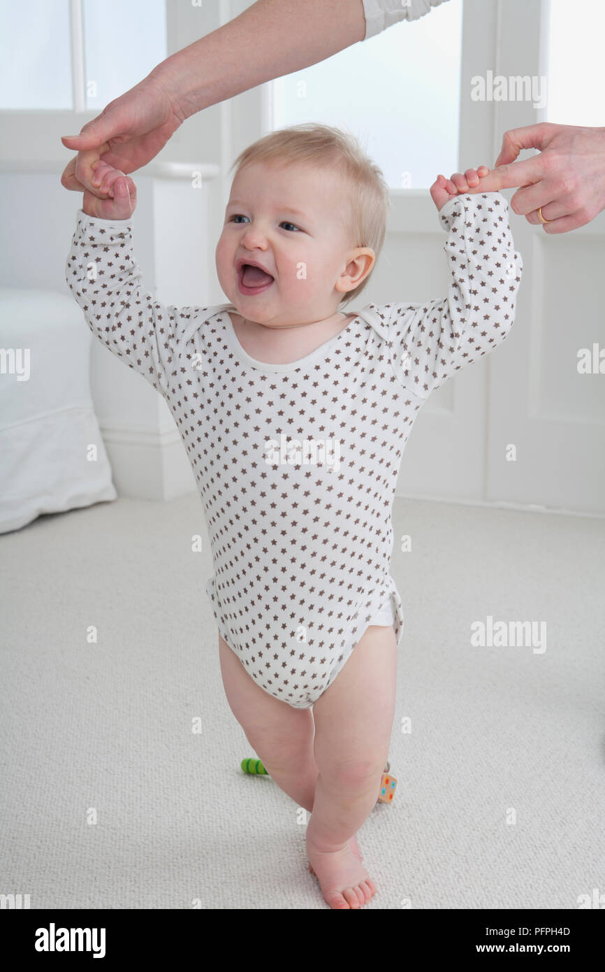 Baby Boy von den Händen seiner Mutter unterstützte, als er lernt, auf dem Teppich zu laufen Stockfoto