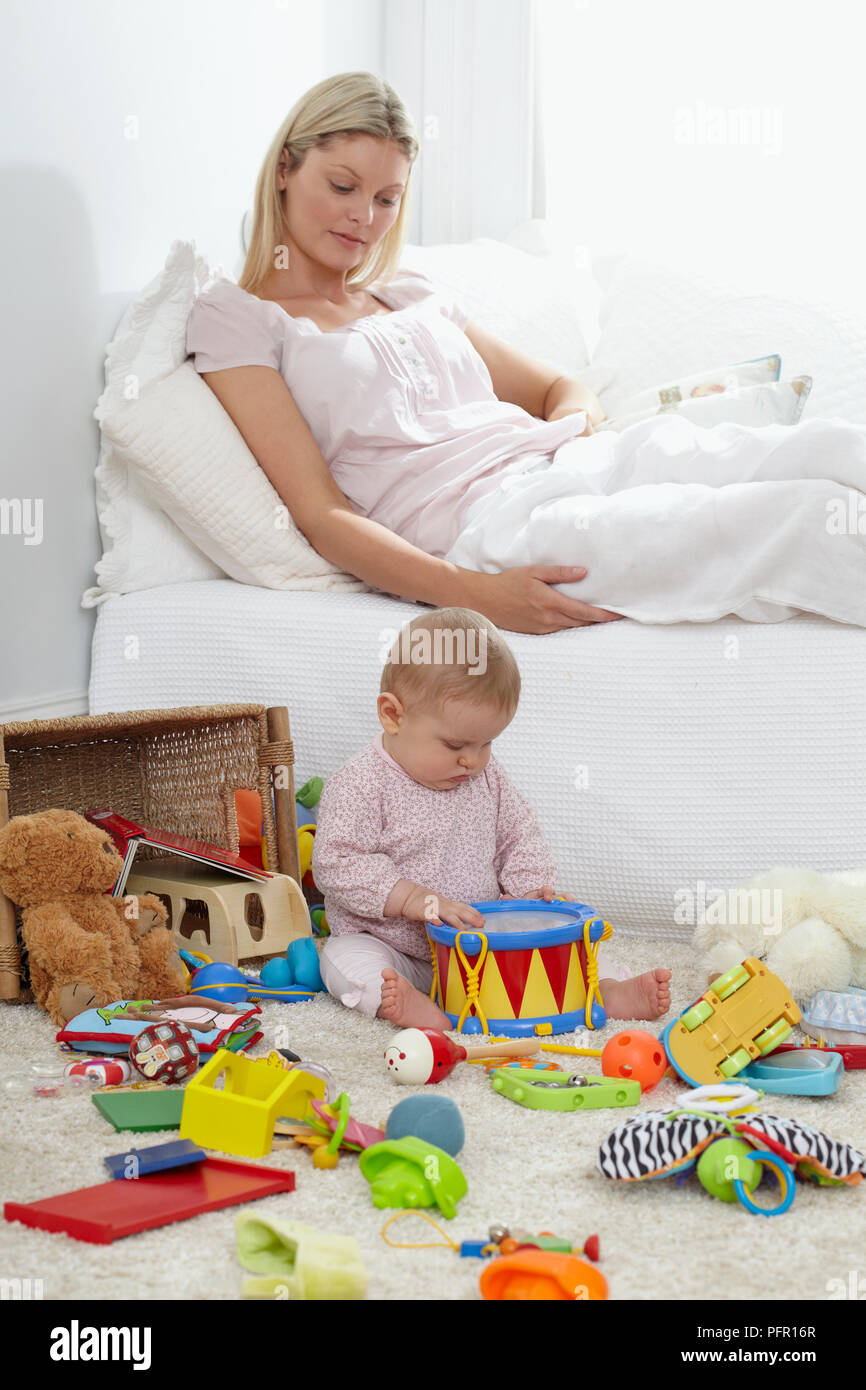 Baby (6 Monate) sitzt auf dem Boden, die von Spielzeug umgeben, Frau hinter ihr sitzt auf einem Sofa Stockfoto